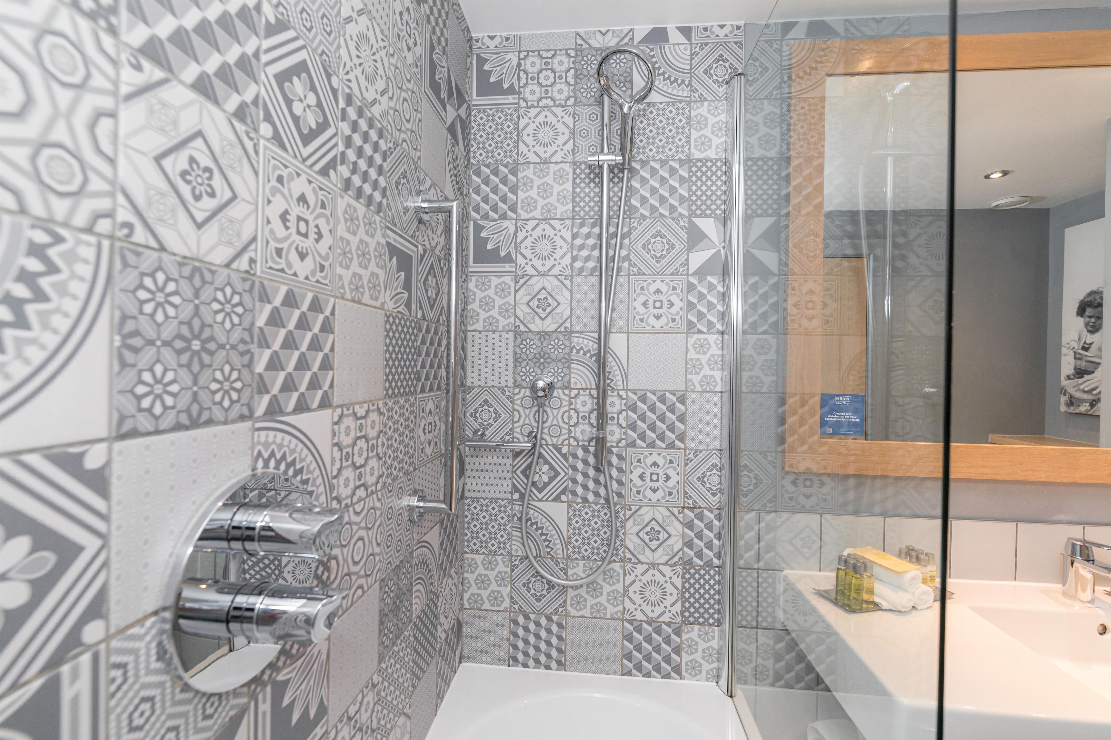 A bathroom with patterned tiles, a glass shower enclosure, a white sink, and a mirror with a wooden frame.