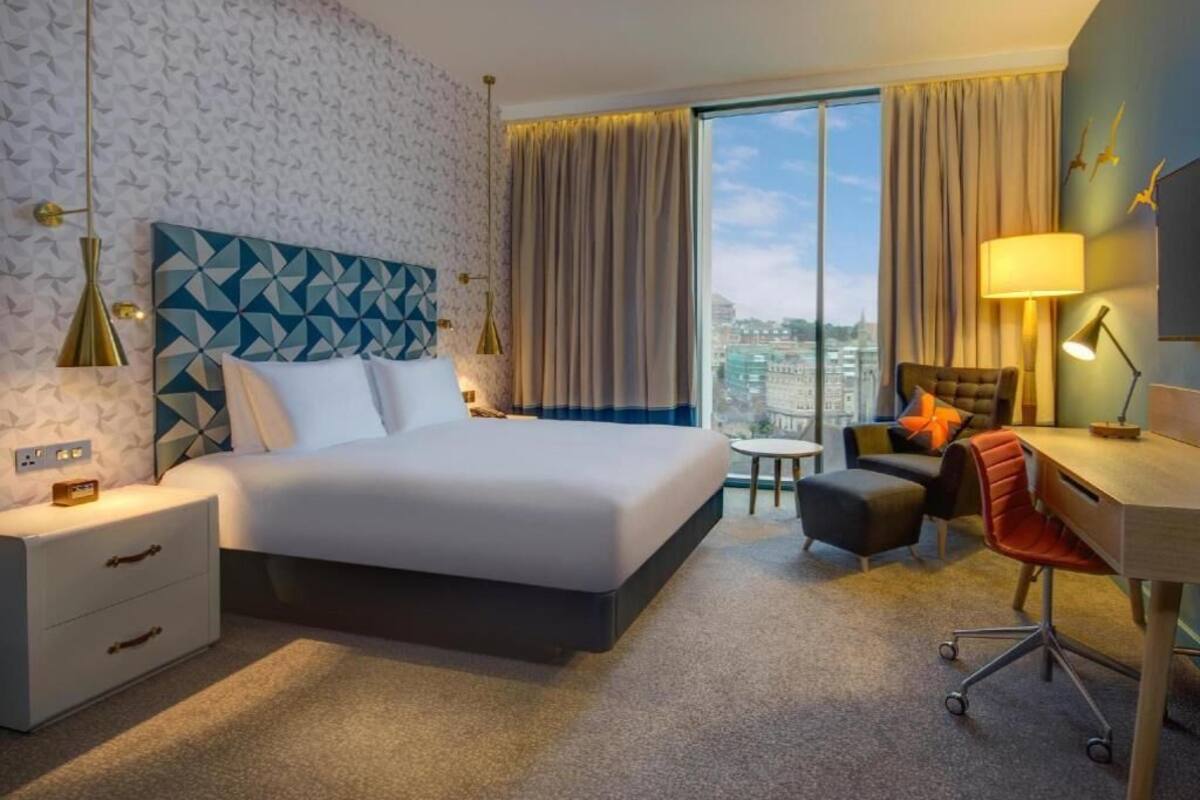 A hotel room with a large bed, a desk, a chair, a nightstand, and a view of the city.