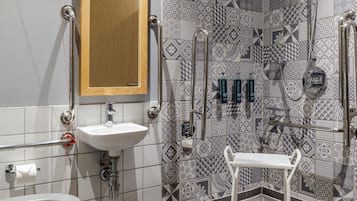 Un baño con lavamanos blanco, espejo, inodoro y zona de ducha con barras de apoyo.