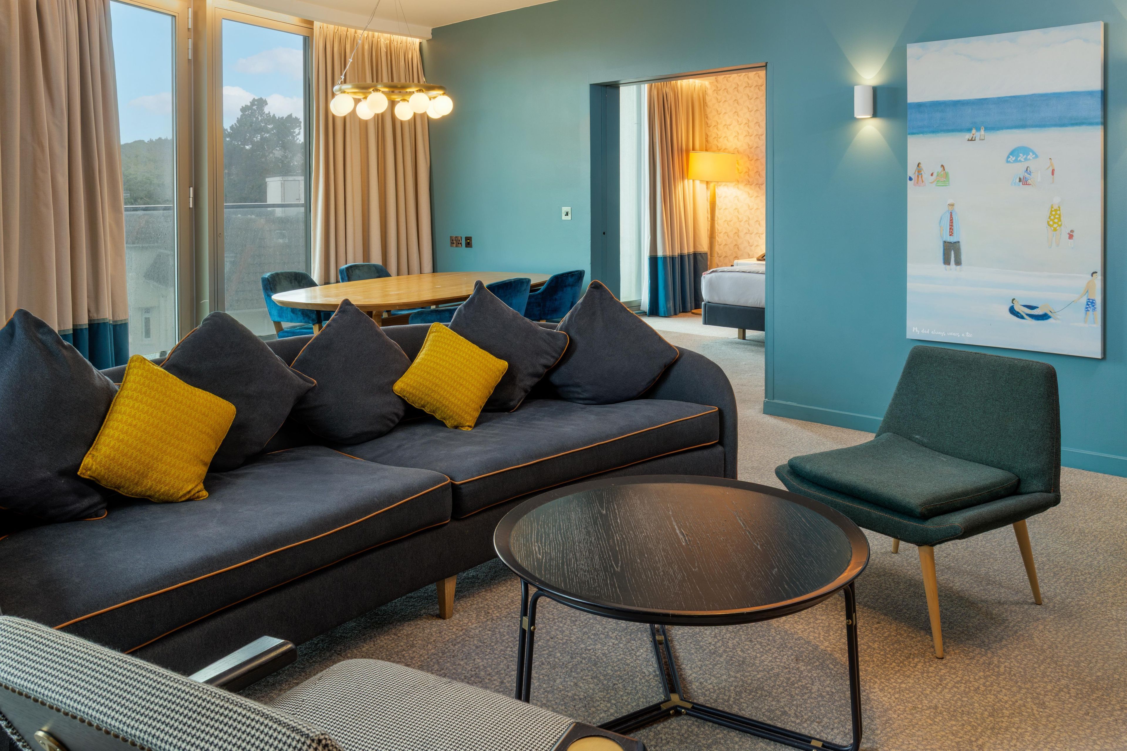 Een moderne hotelkamer met een donkergrijze bank, een ronde tafel en een groene fauteuil.