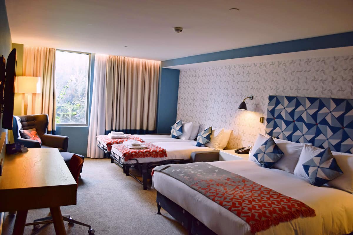 A hotel room with two beds, a desk, a chair, a lamp, and a view of greenery outside.