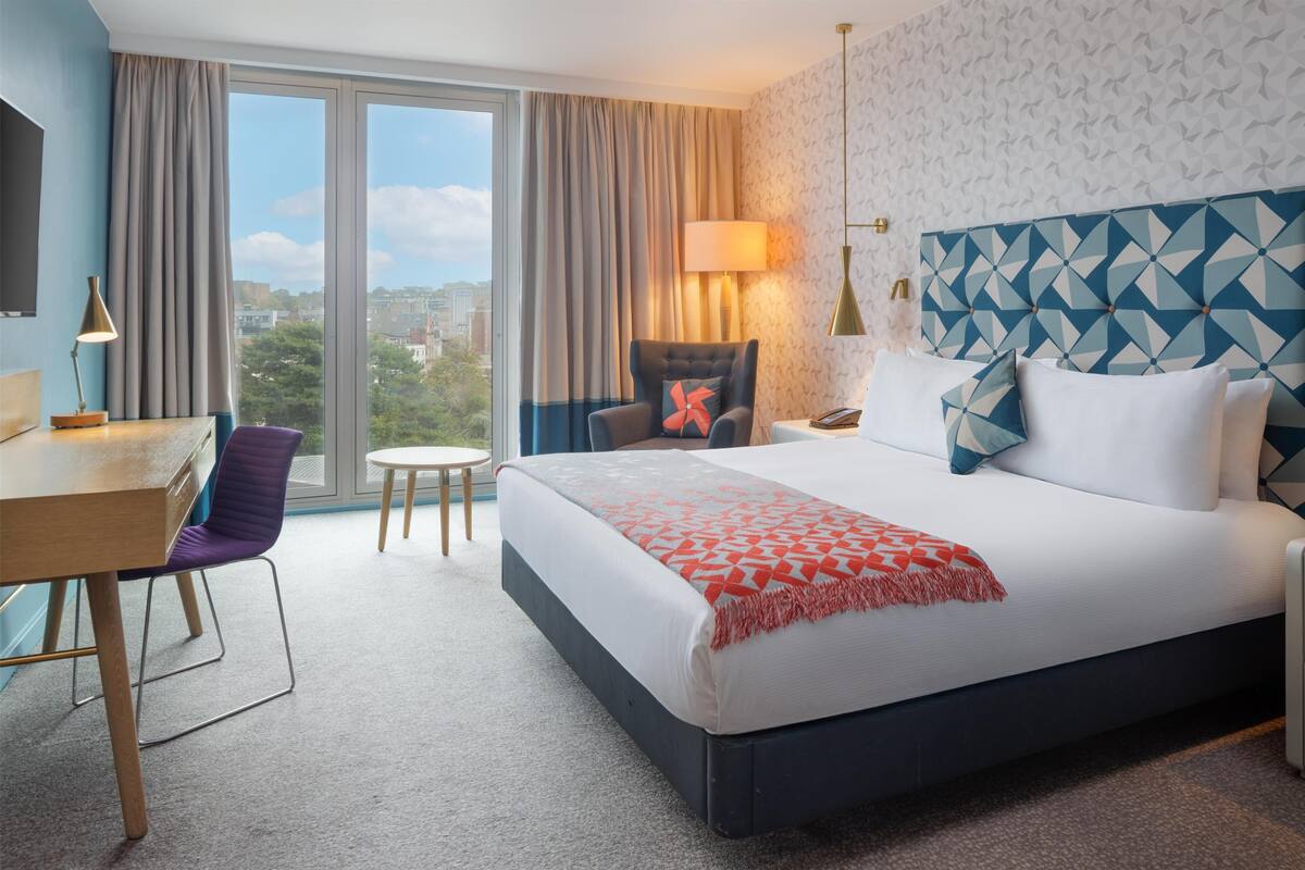 A hotel room with a large bed, a desk, a chair, a lamp, and a view of the city through large windows.
