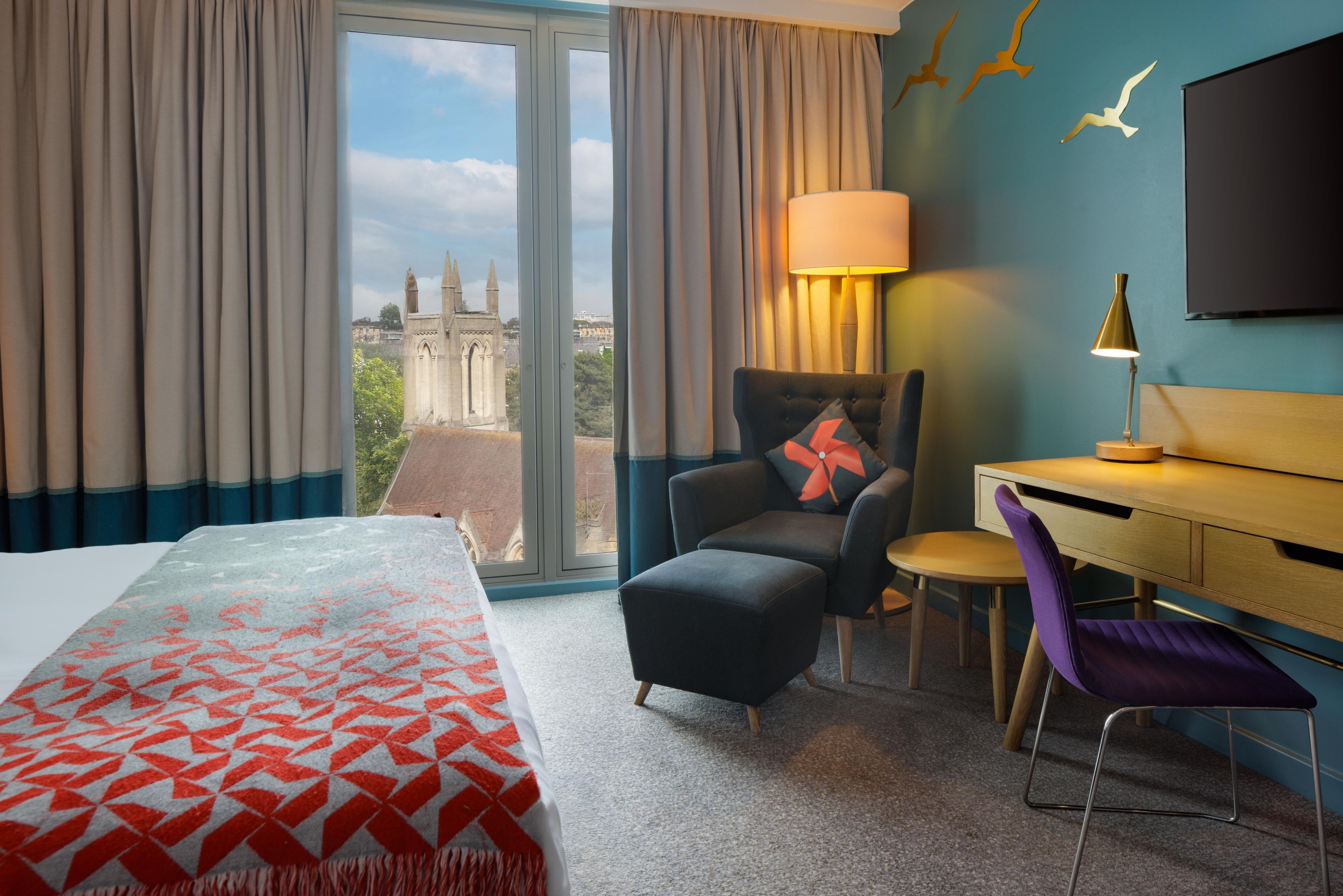 A hotel room with a bed, a desk, a chair, a lamp, and a view of a building through the window.