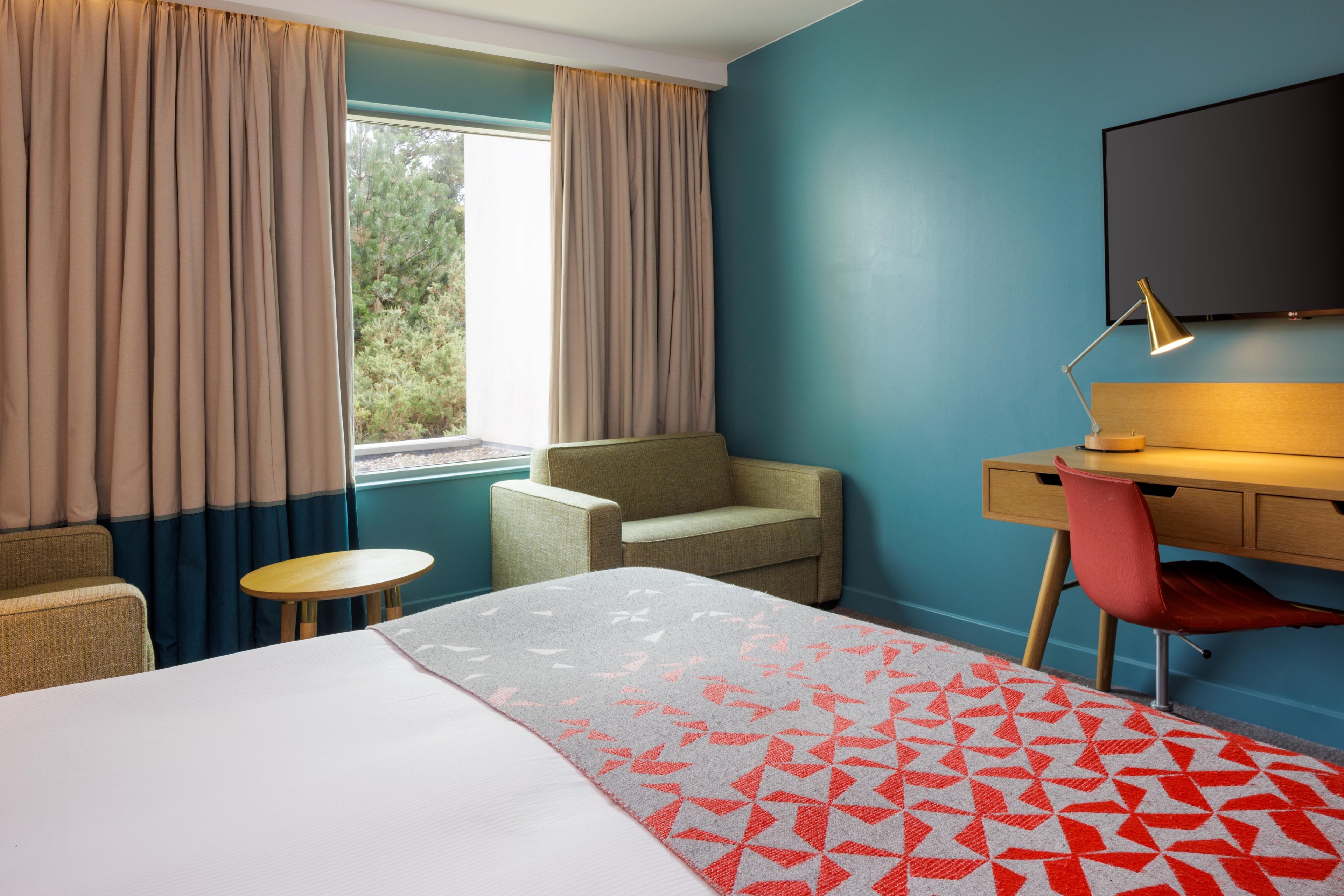 A hotel room with a bed, a desk, a chair, a small table, and a TV.