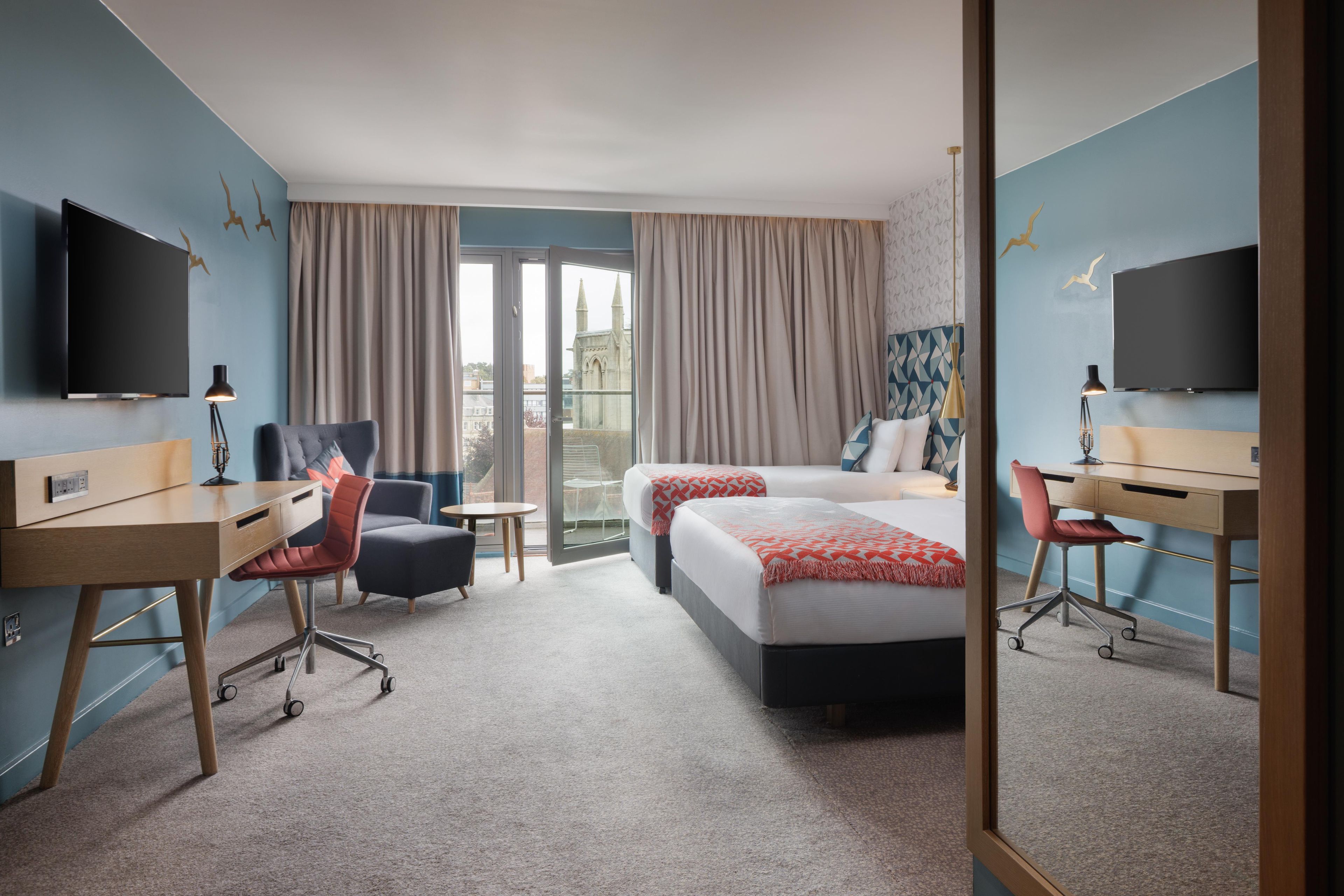 Une chambre d’hôtel avec un lit, un bureau, une chaise, une télévision et un balcon avec des rideaux.