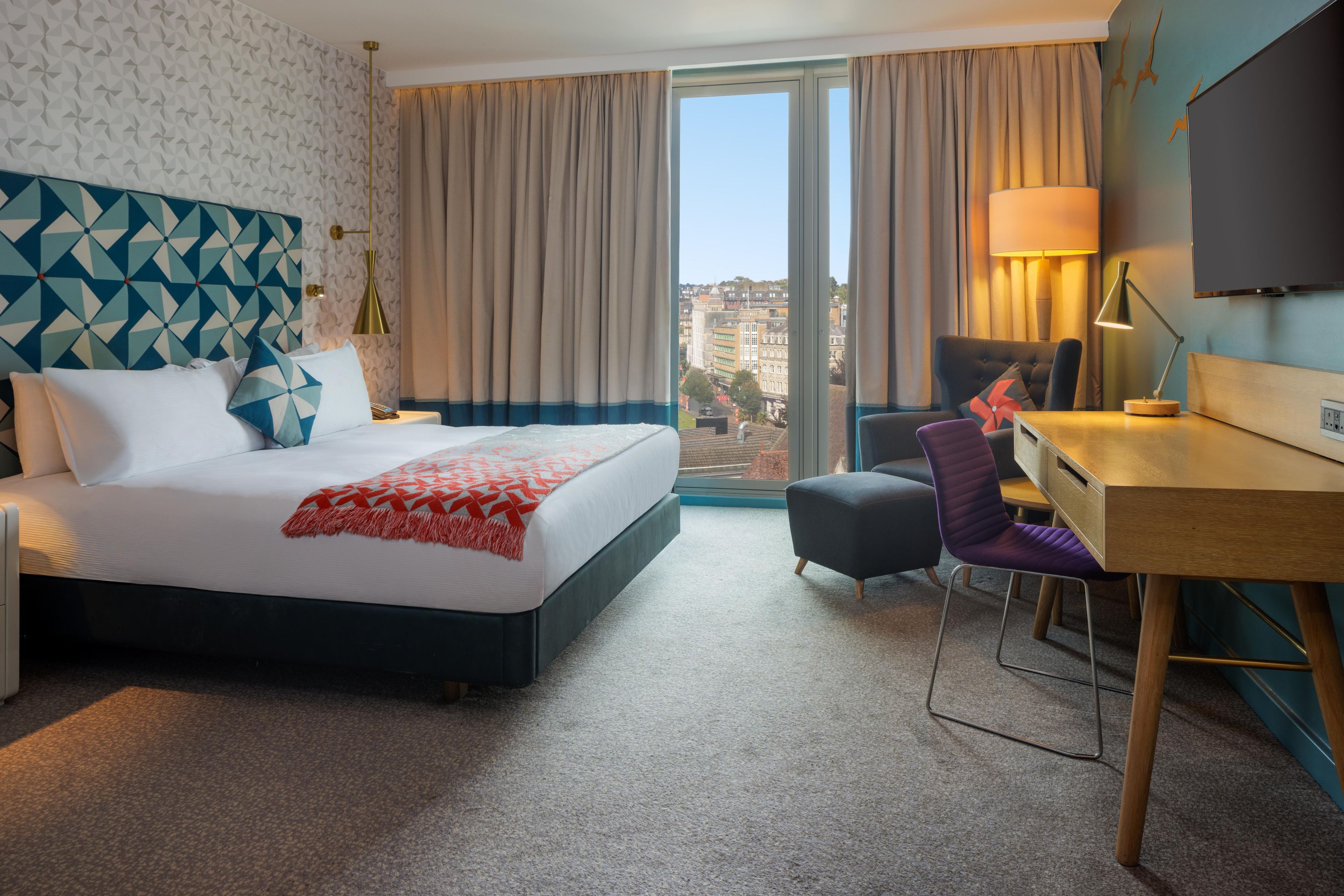 A hotel room with a bed, a desk, a chair, and a view of the city.