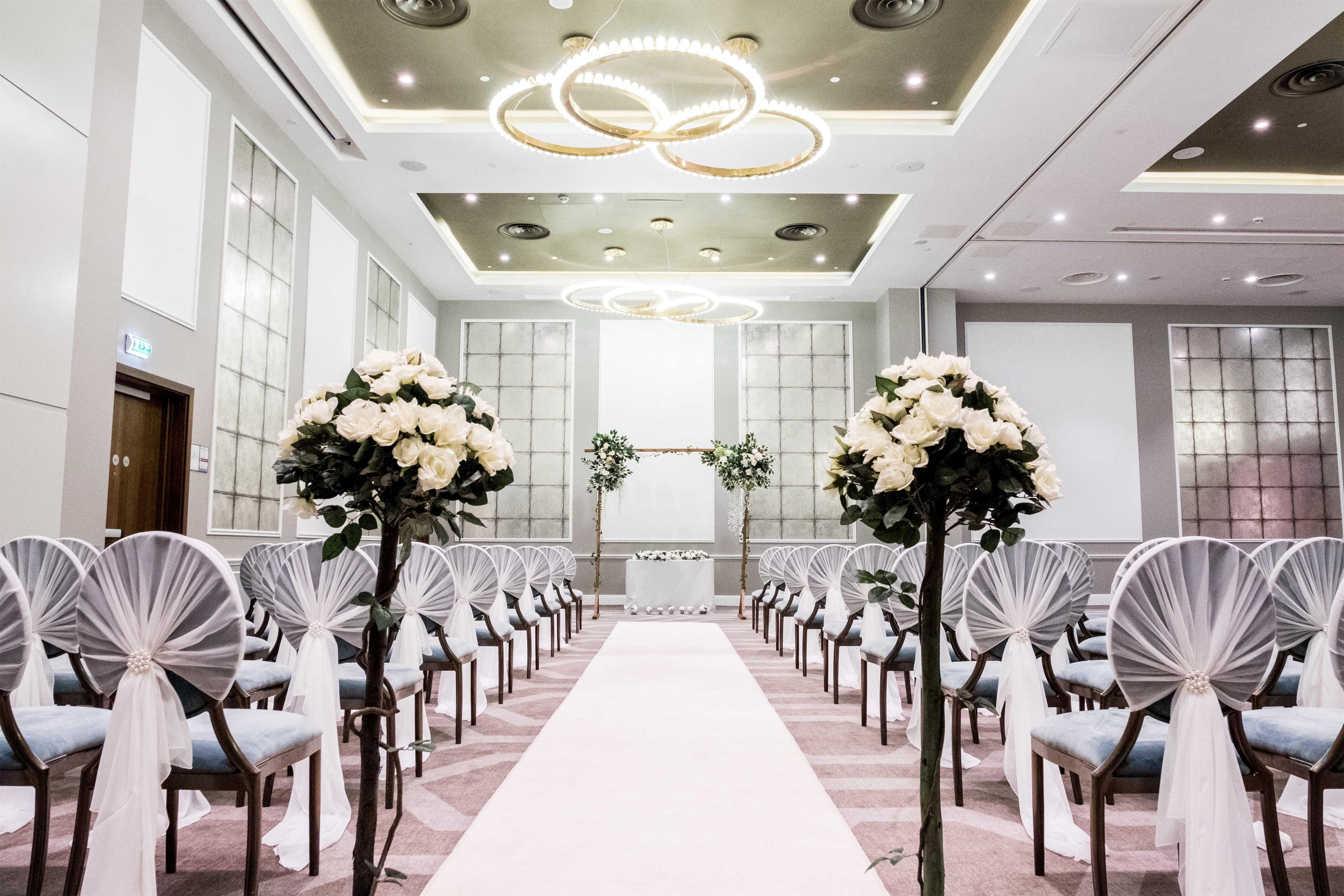 A hotel chapel with a white aisle, pews with white and blue cushions, and a floral arrangement at the altar.