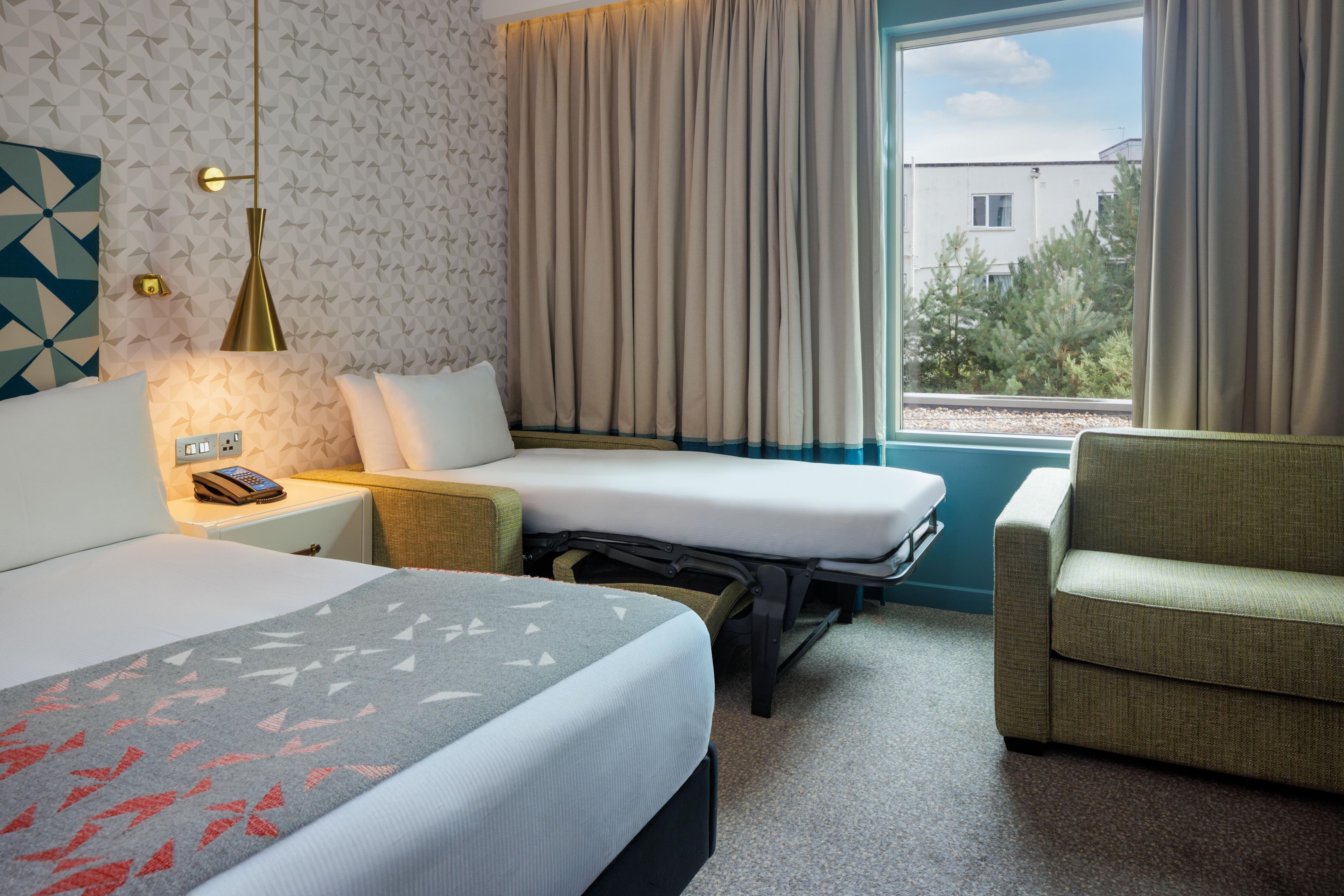 A hotel room with a bed, bedside table, lamp, chair, and a window with curtains.