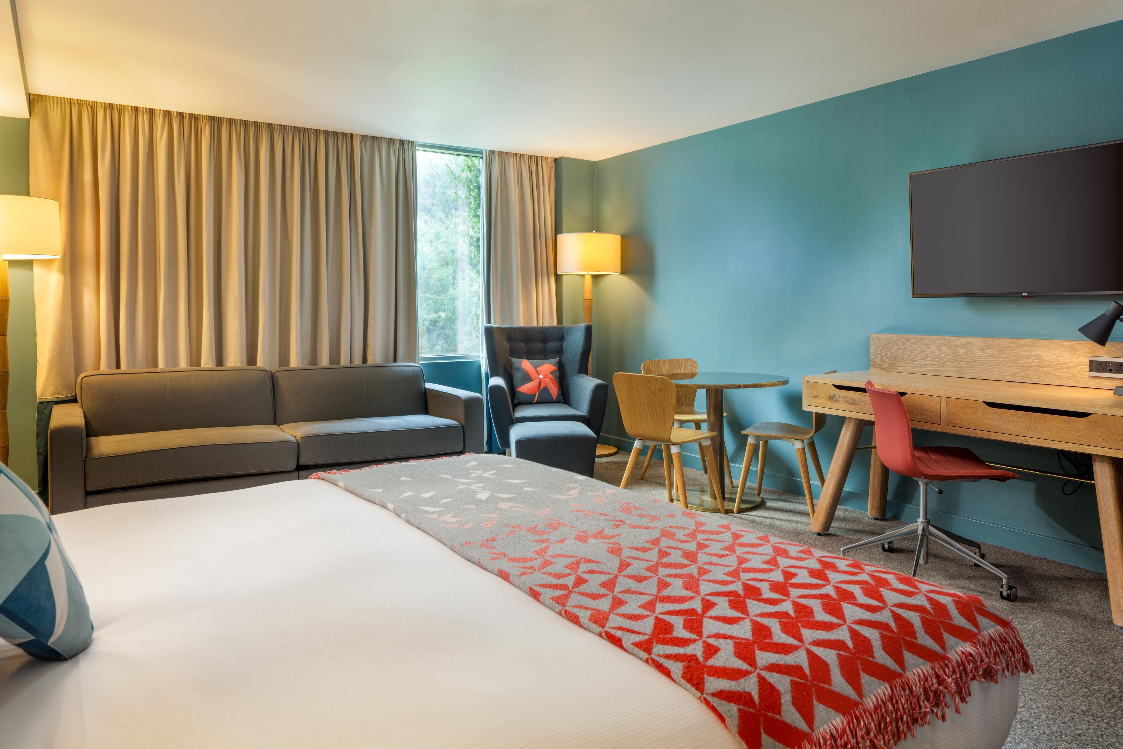 A hotel room with a bed, a sofa, a desk, a chair, a TV, and a window with curtains.