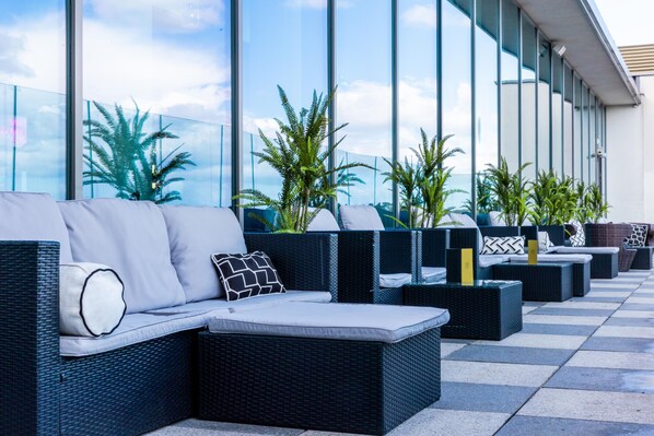 Terraza exterior con mobiliario de mimbre y cojines, plantas en macetas y fachada de vidrio.
