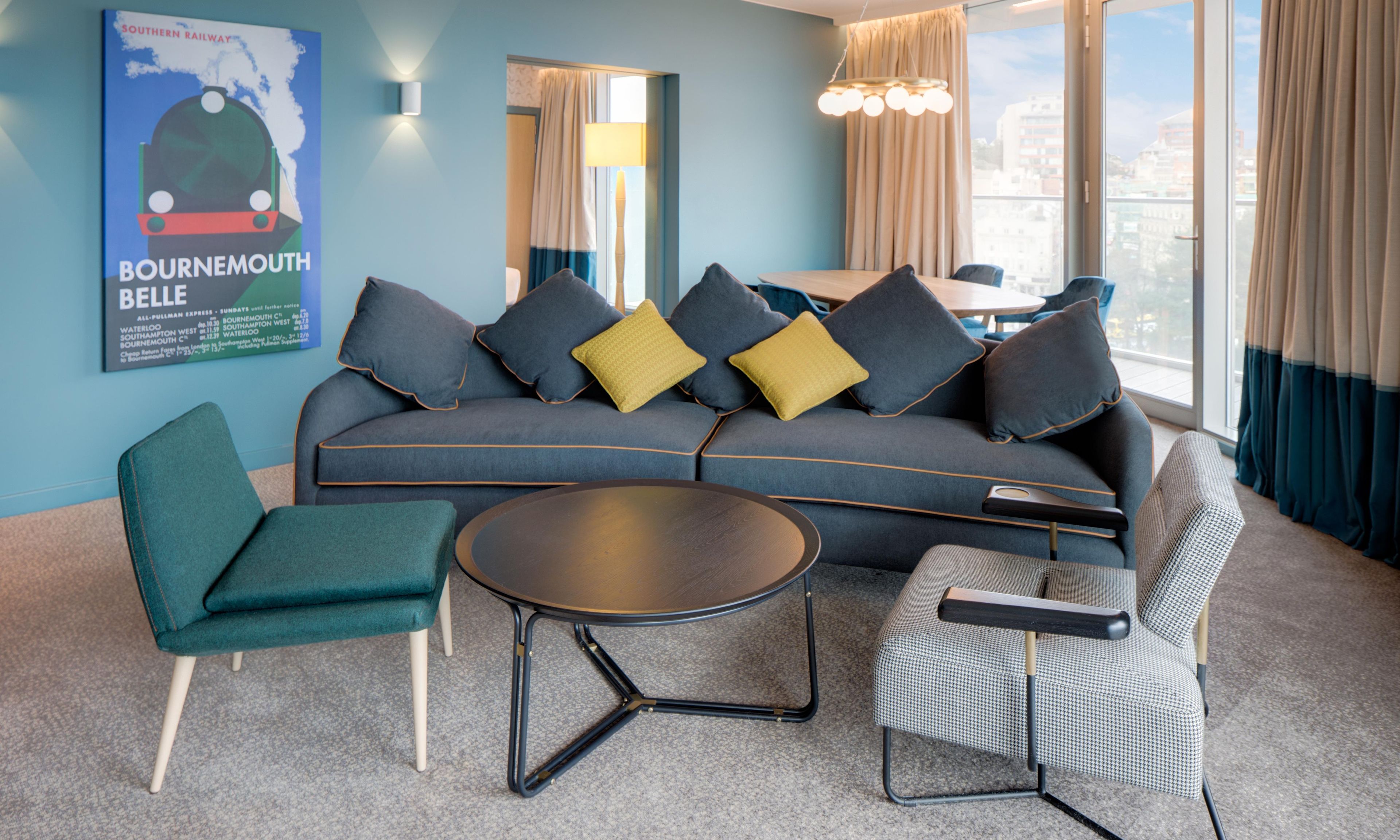 A modern hotel room with a sofa, two armchairs, a coffee table, and a poster on the wall.