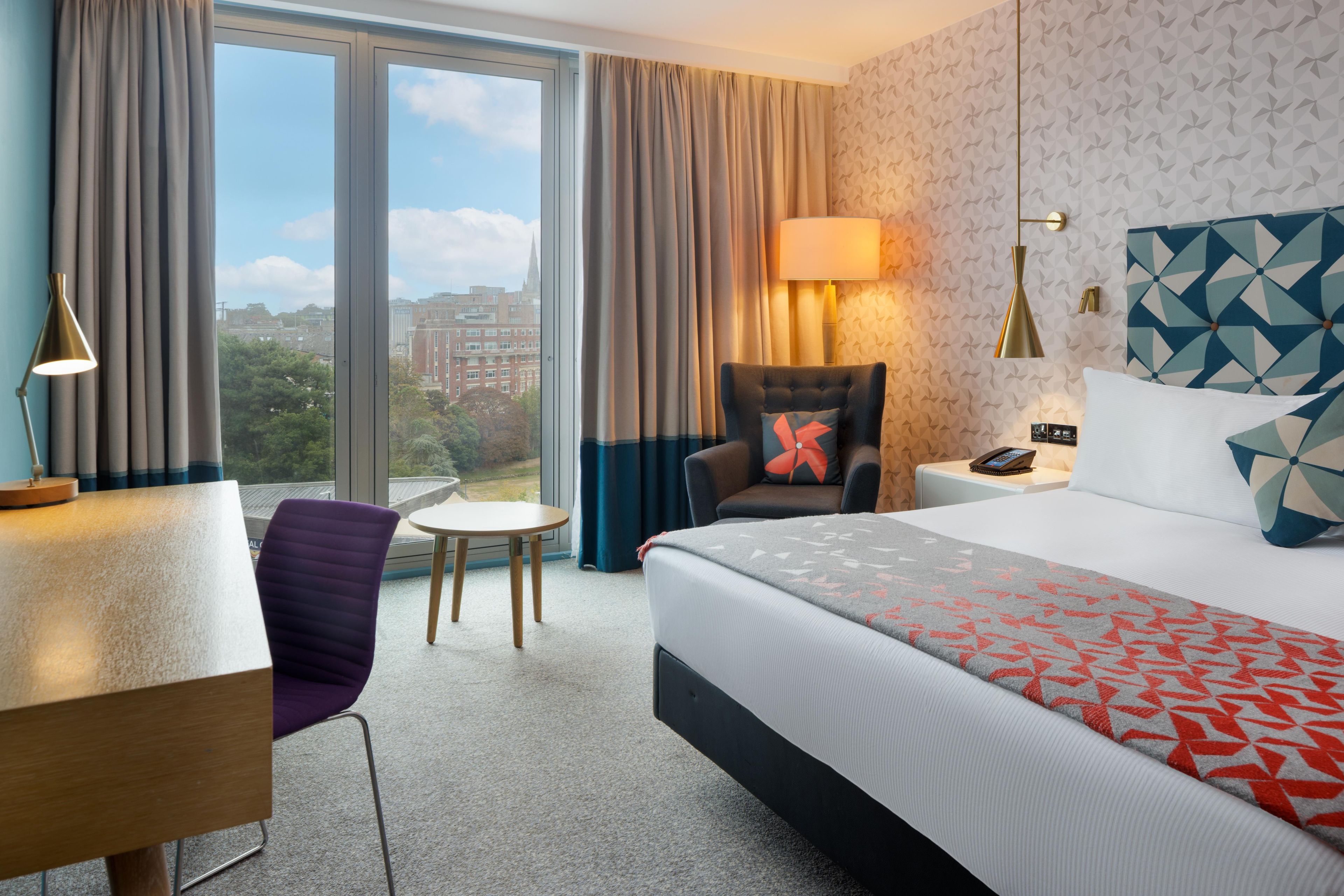 A hotel room with a bed, a desk, a chair, a lamp, and a view of the city.