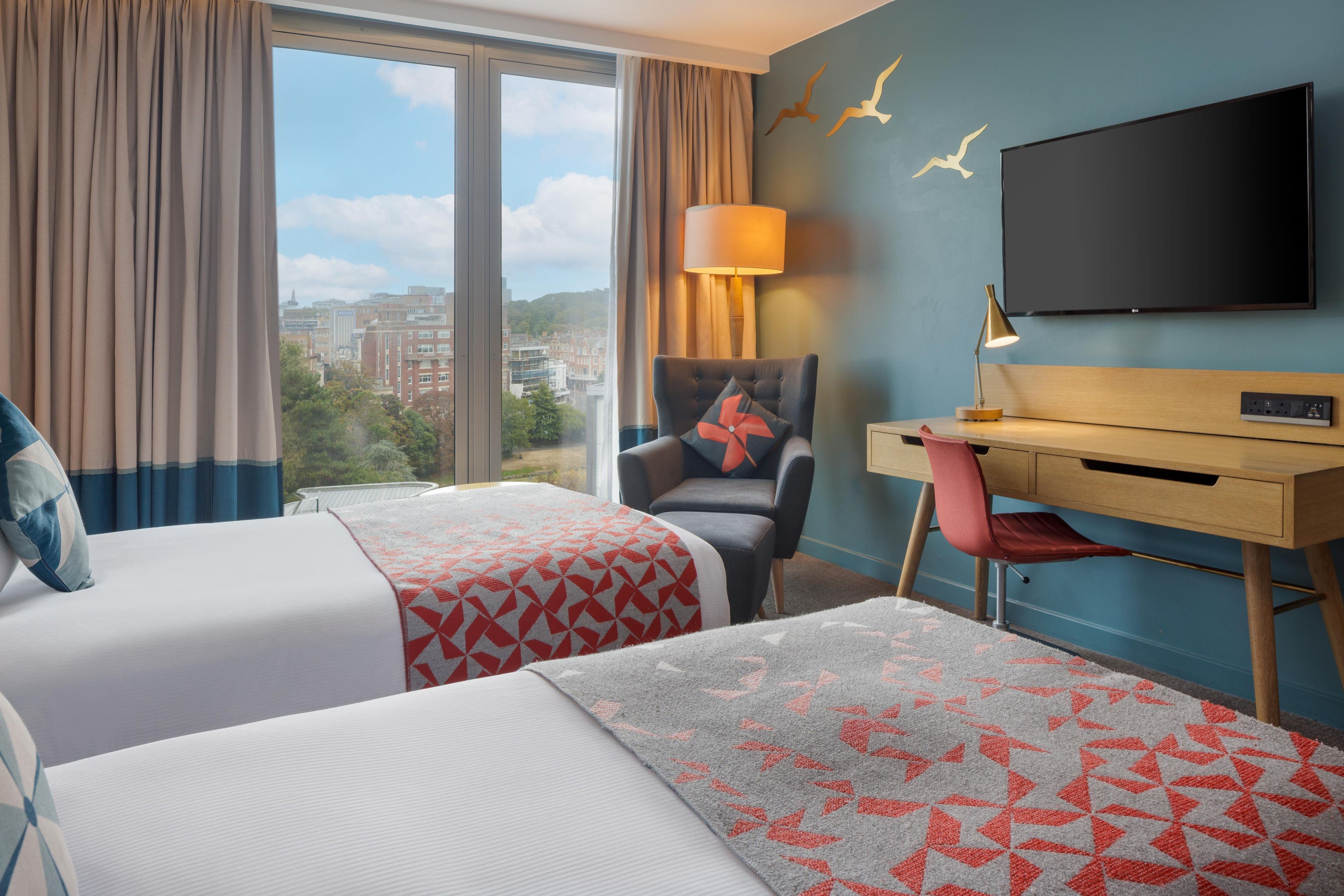 Een hotelkamer met twee bedden, een bureau, een stoel, een televisie en uitzicht op de stad.