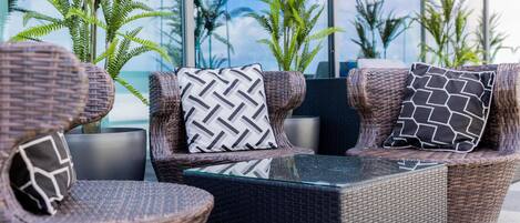 Mobiliario de mimbre exterior con cojines estampados, una mesa con superficie de vidrio y plantas en macetas.
