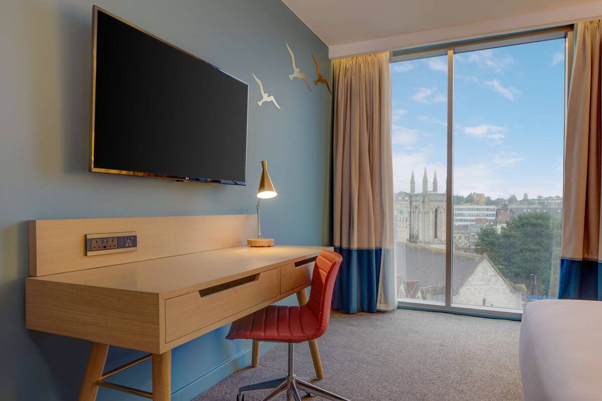 A hotel room with a wooden desk, a red chair, a flat-screen TV, and a large window offering a city view.