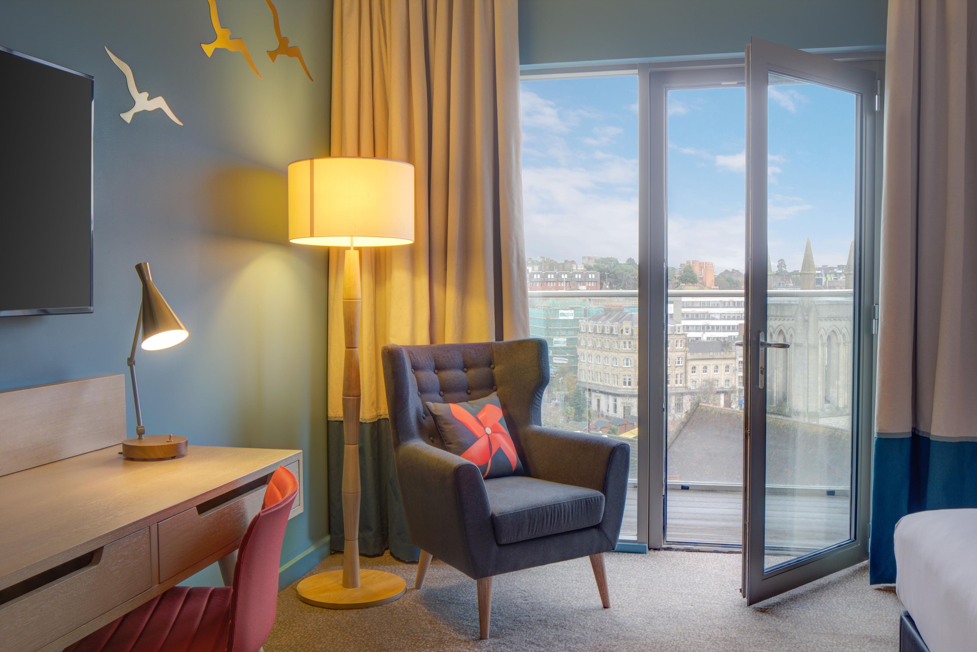 A hotel room with a desk, chair, lamp, and a view of the city through a glass door.