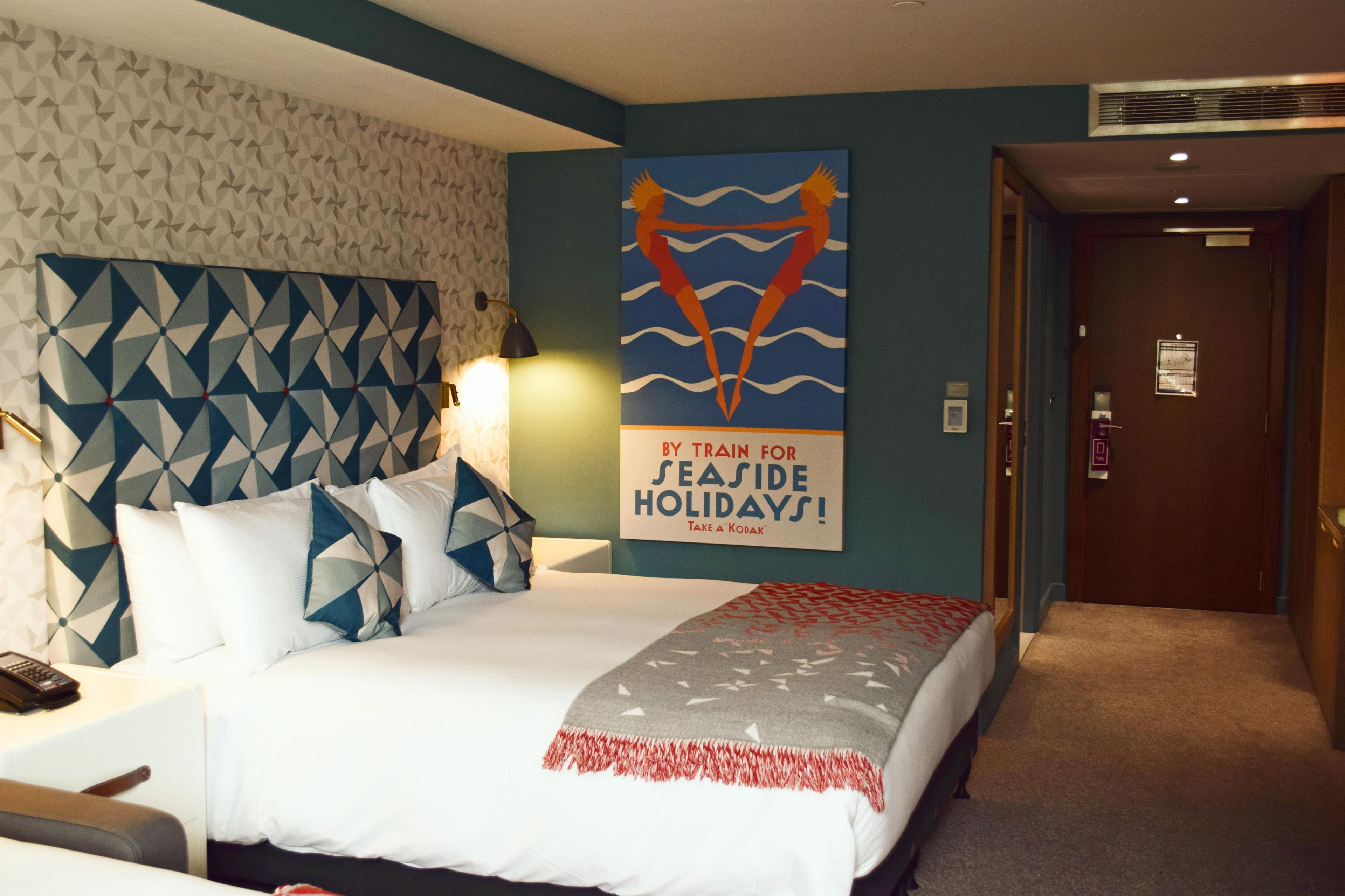 A hotel room with a bed, pillows, a blanket, a wall with a geometric pattern, and a poster advertising seaside holidays.