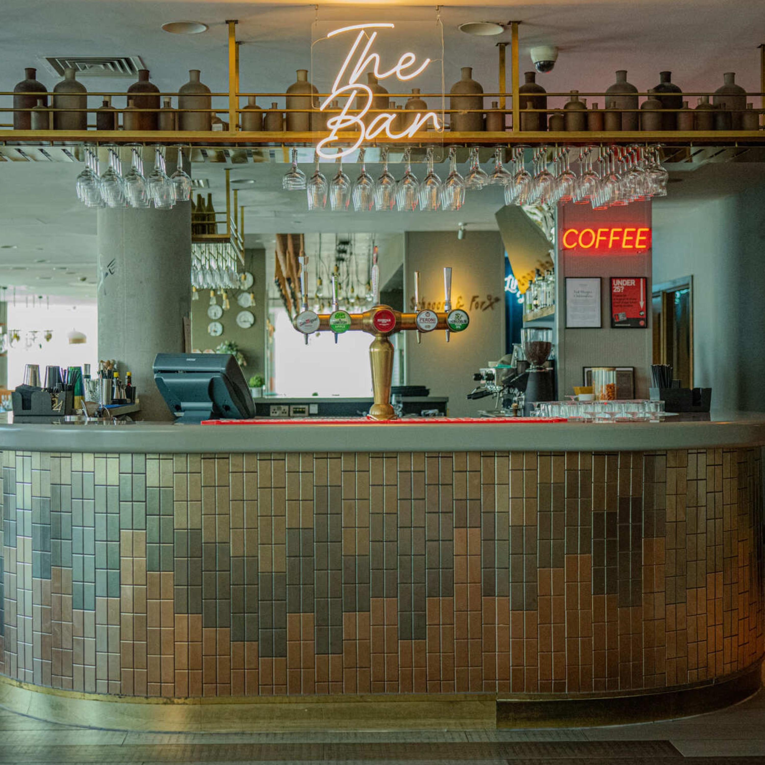 Eine Bar mit einer Theke, einem leuchtenden Neon-Schild mit der Aufschrift 'The Bar' und einem Schild für Kaffee.