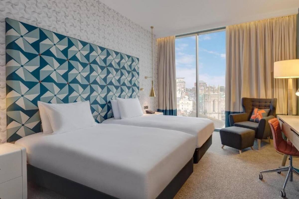 A hotel room with a large bed, a desk, a chair, and a view of the city.