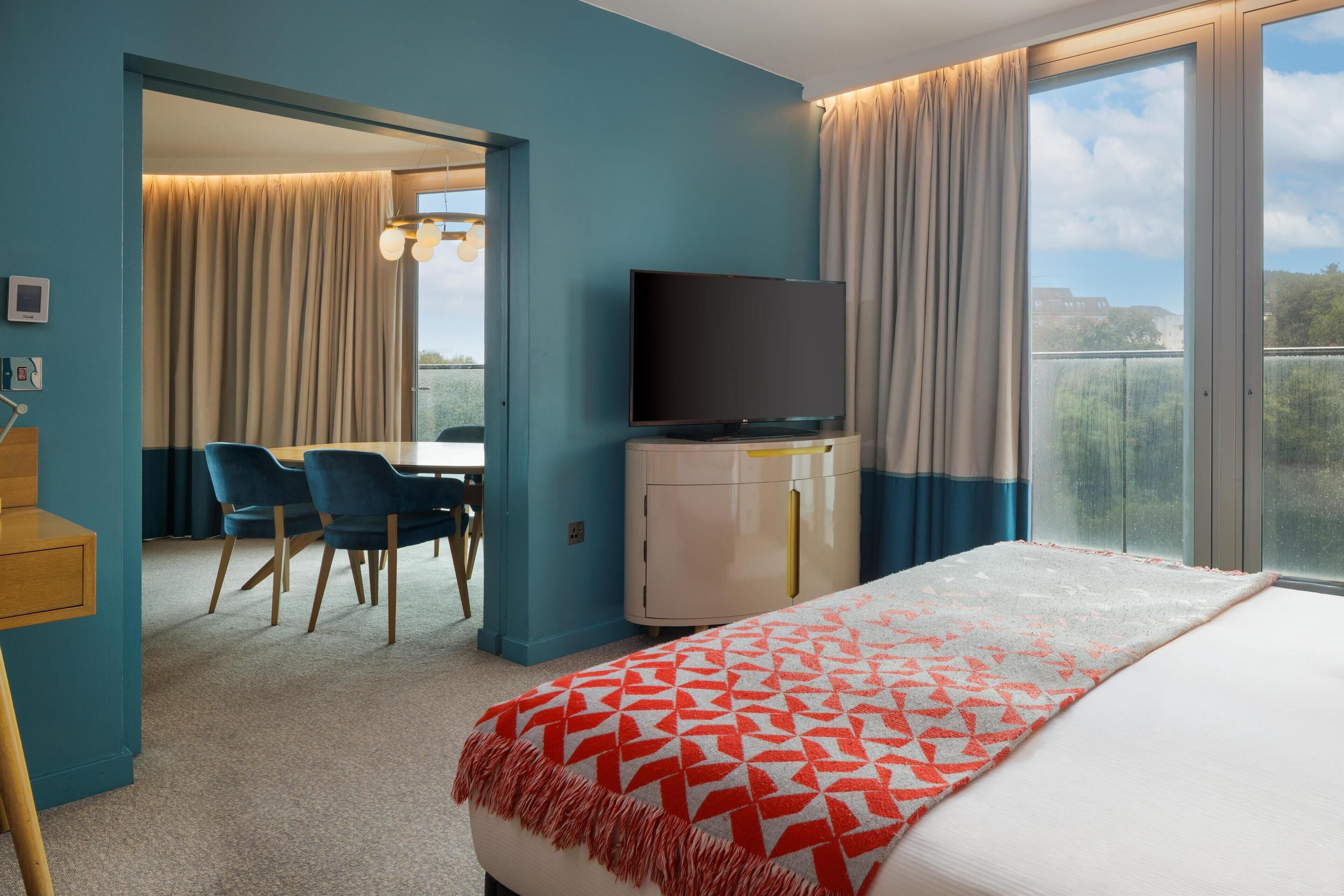 Habitación de hotel con cama, televisión, escritorio y sillas, y vista al exterior.