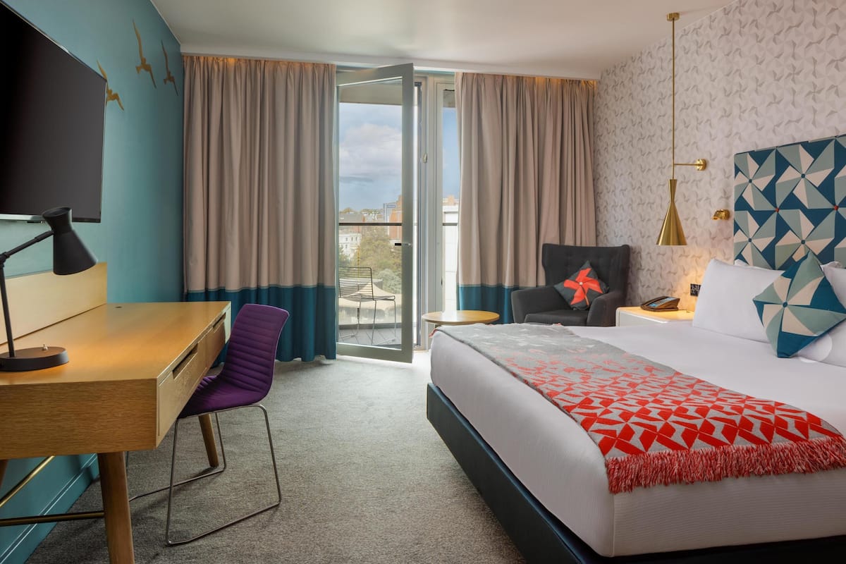 A hotel room with a bed, a desk, a chair, a TV, and a balcony with curtains.