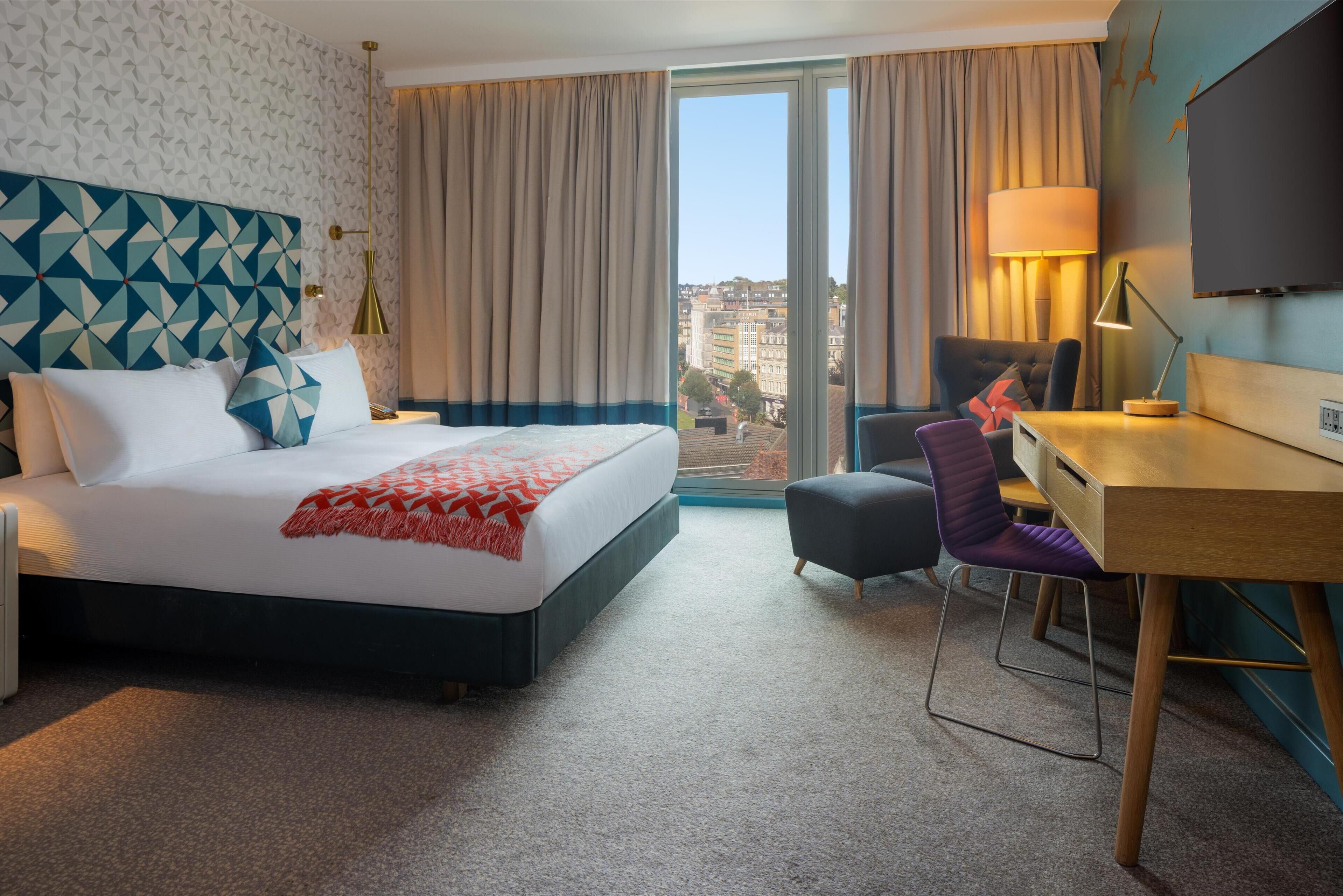 A hotel room with a large bed, a desk, a chair, and a view of the city through the window.