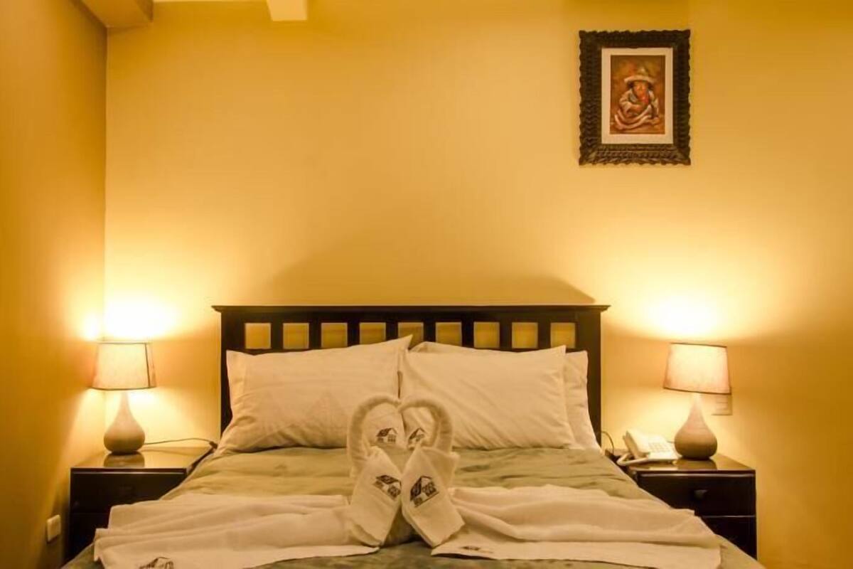 A neatly made bed with white linens and a towel with a house design, two bedside lamps, and a framed picture on the wall.