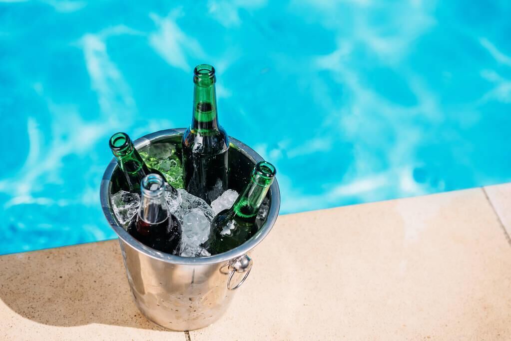 Un cubo de metal lleno de hielo y tres botellas verdes, junto a una piscina.