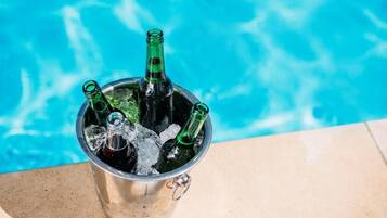 Un cubo de metal lleno de hielo y tres botellas verdes, junto a una piscina.