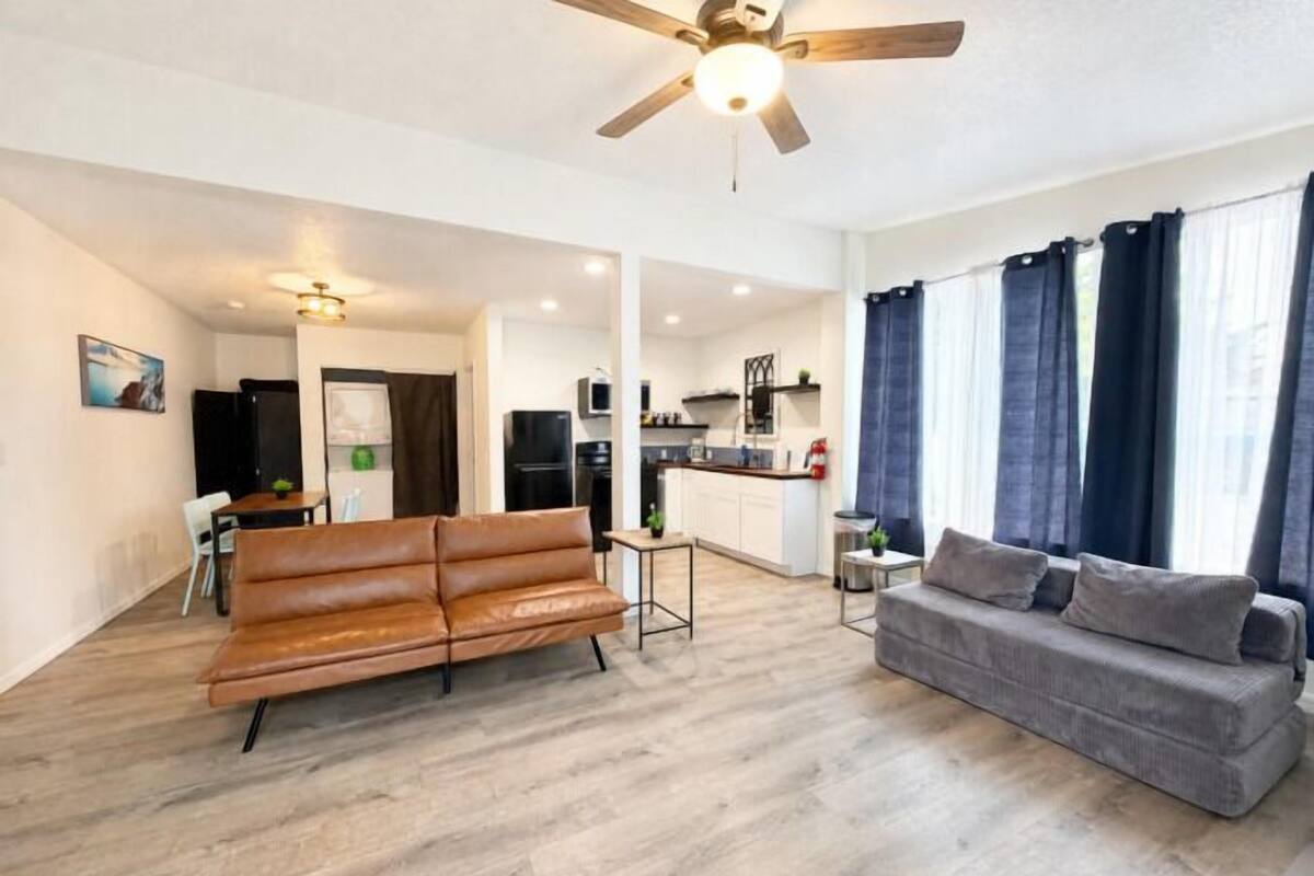 A modern living room with a sofa and a couch, a ceiling fan, and a kitchen area in the background.