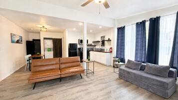 A modern living room with a sofa and a couch, a ceiling fan, and a kitchen area in the background.