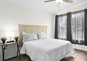 Un dormitorio ordenado con cama, mesita de noche, lámpara y cortinas.