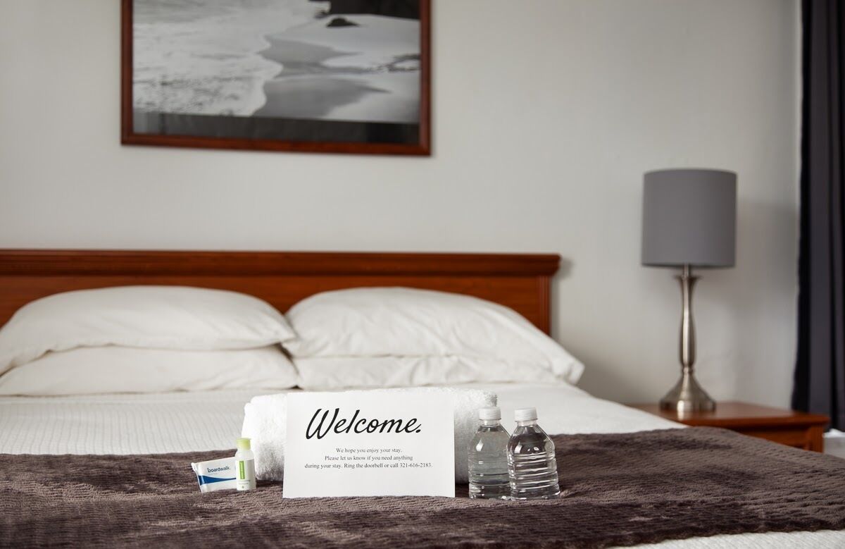 A hotel room with a bed, a welcome sign, a bedside lamp, and a framed picture on the wall.