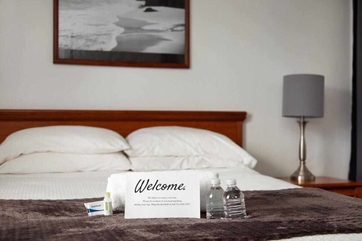 A hotel room with a bed, a welcome sign, a bedside lamp, and a framed picture on the wall.