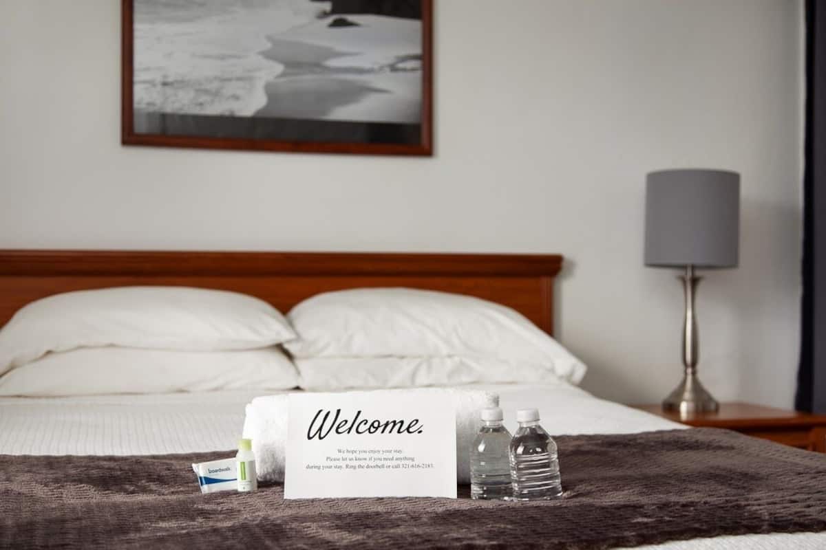 Hotellihuone, jossa on sänky, tervetulokyltti, yöpöytälamppu ja seinällä oleva kehystetty kuva.