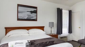 Ein Hotelzimmer mit Bett, Nachttisch mit Lampe, Fenster mit Vorhängen, einer Tür und einem gerahmten Bild an der Wand.