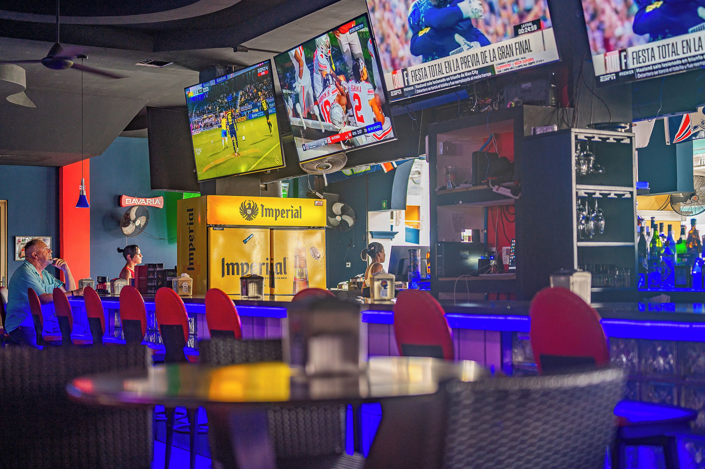 A bar area with multiple screens displaying sports events, a Bavaria sign, and an Imperial sign.