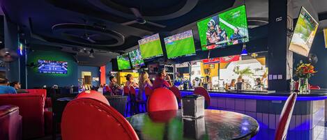 A bar area with multiple screens displaying sports events, red seating, and a counter with a bartender.