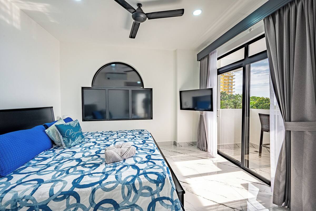 Habitación de hotel moderna con cama, ventilador de techo, televisión y balcón con vistas a un edificio y zonas verdes.