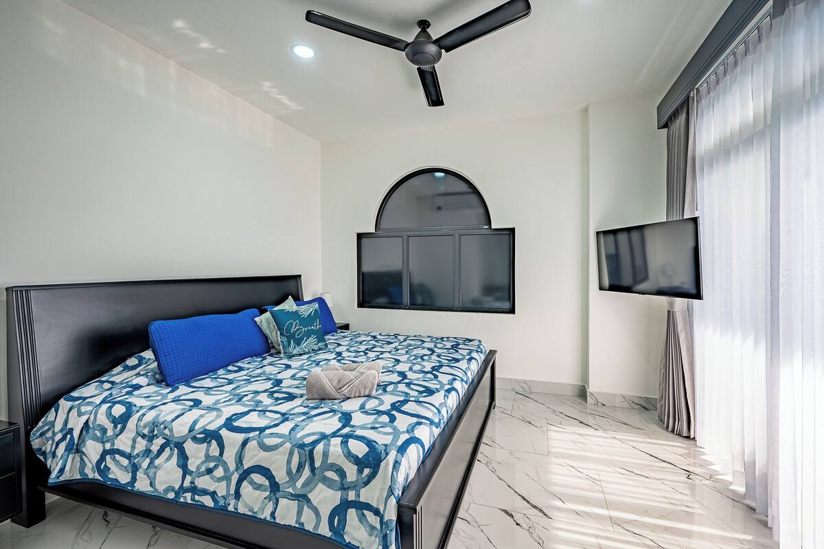 Un dormitorio con cama, ventilador de techo, televisor y ventana con cortinas.
