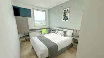 Quarto moderno com uma cama grande, uma escrivaninha e uma cadeira. Há uma janela com vista para edifícios e um quadro na parede.