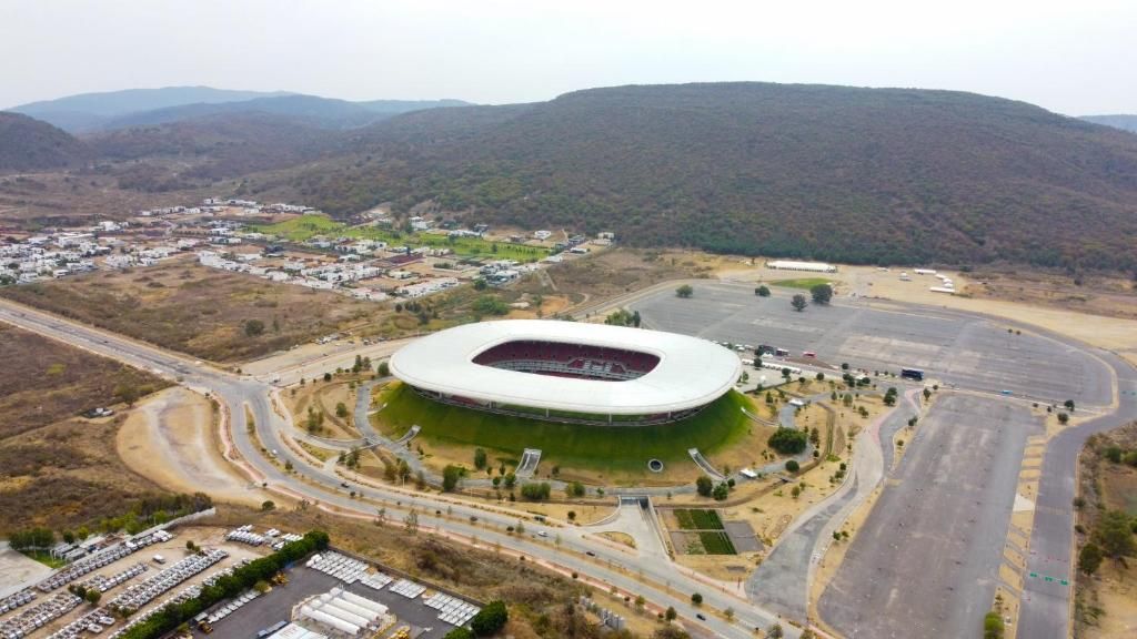 Un estadio con un amplio campo central rodeado de gradas, caminos y zonas de estacionamiento.
