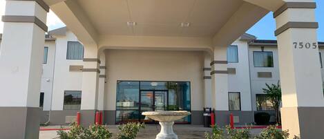 A modern building entrance with a covered walkway, a central fountain, and a number '7505' on the right pillar.