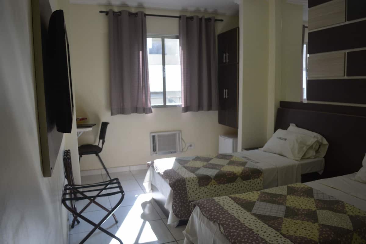 Ein Schlafzimmer mit zwei Betten, einem Sessel, einem Fenster mit Vorhängen und einem an der Wand befestigten Fernseher.