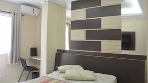 Habitació d'hotel amb un llit, una taula de treball i una cadira. Hi ha una finestra amb cortines, un aire condicionat i una televisió penjada a la paret.