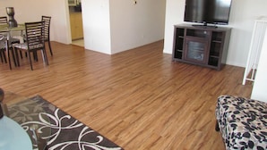 Una sala de estar con piso de madera, un televisor sobre un mueble bajo, una zona de comedor con sillas y una mesa, y una cocina al fondo.