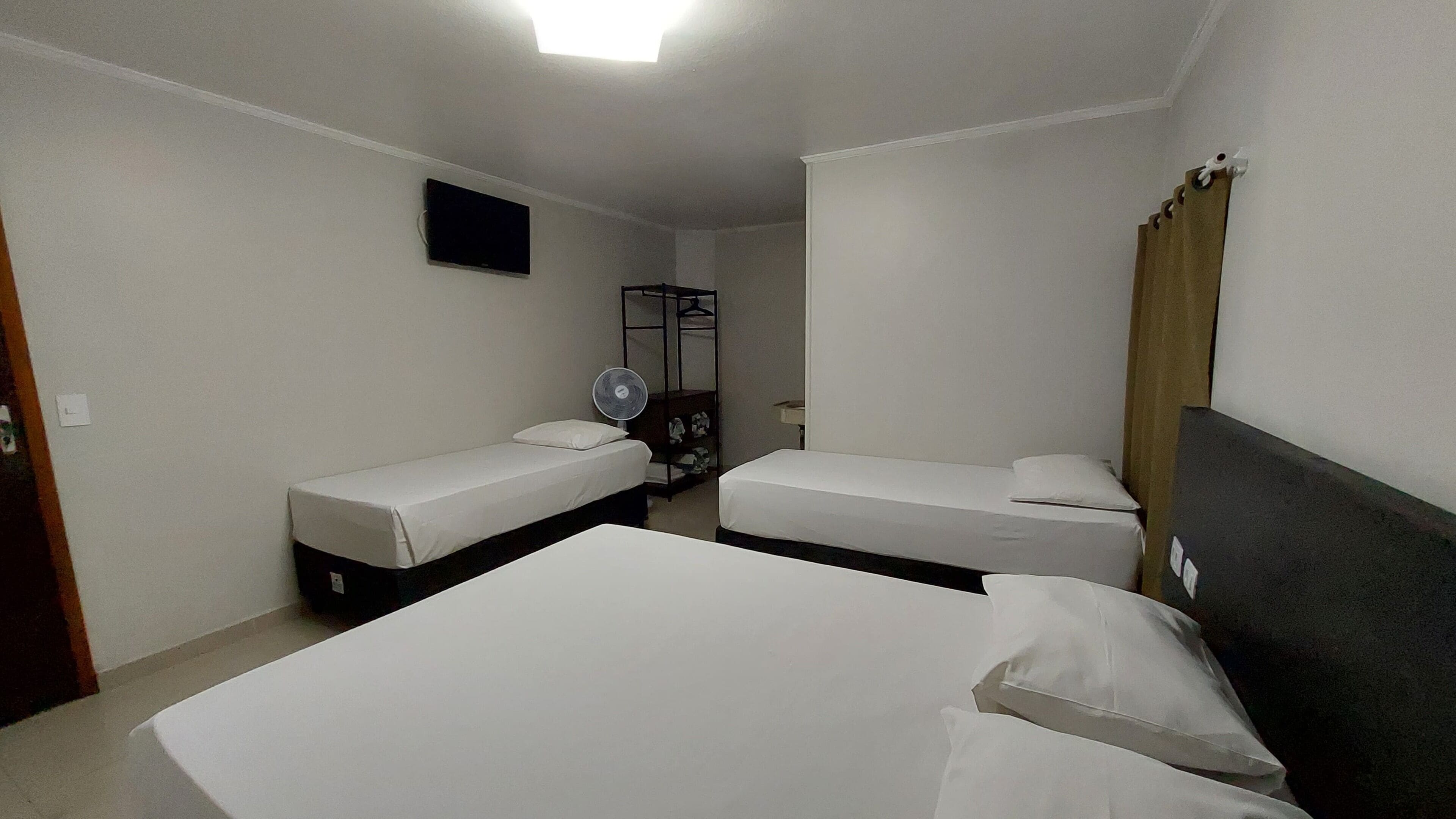 A hotel room with two single beds, a flat-screen TV mounted on the wall, a fan, and a wooden shelf with clothes.