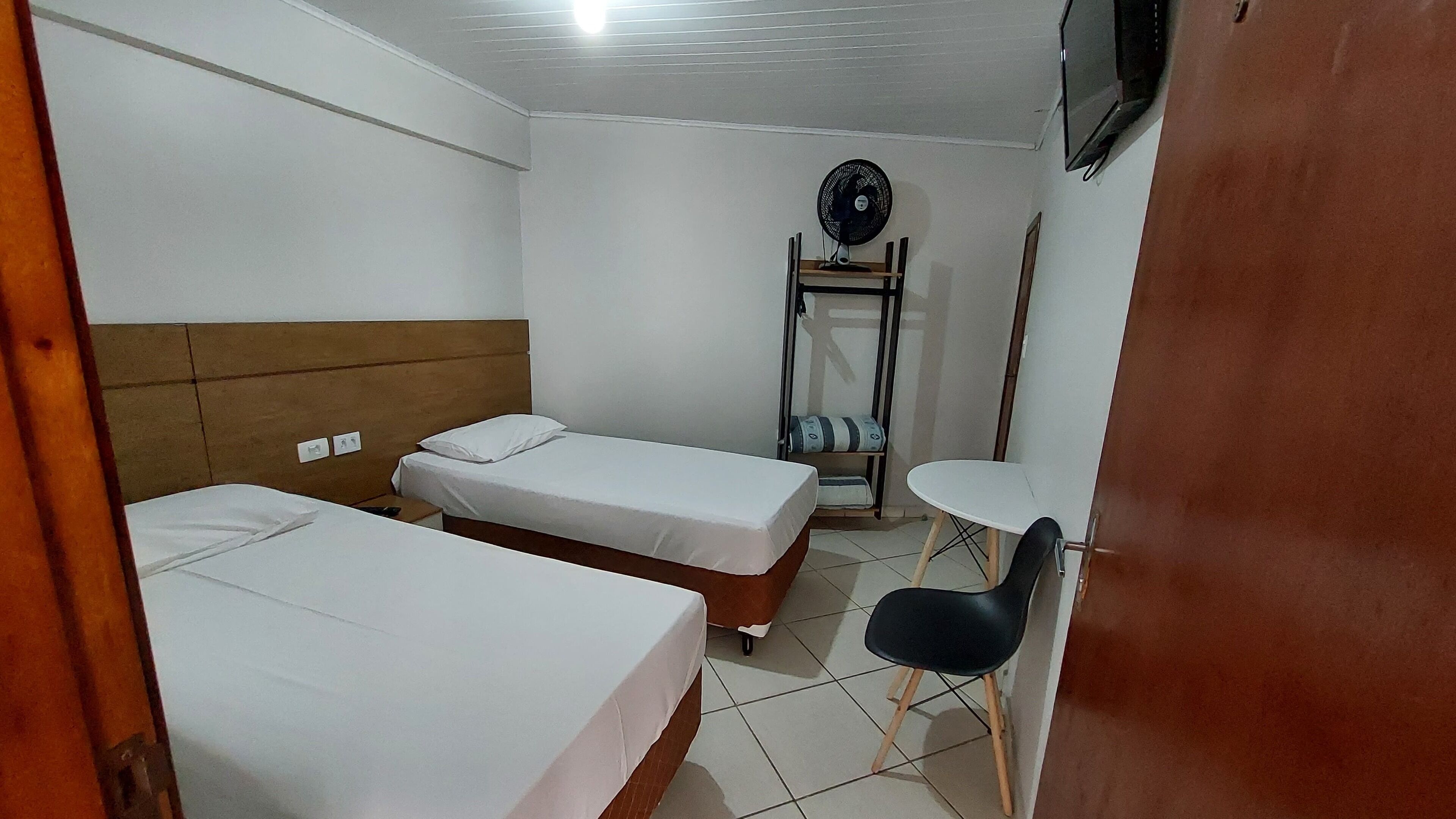 Une petite chambre propre, avec deux lits simples, une petite table, une chaise, un ventilateur et un téléviseur fixé au mur.