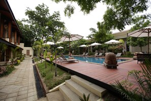 A pool area with lounge chairs and umbrellas, surrounded by greenery and a building with a tiled roof.