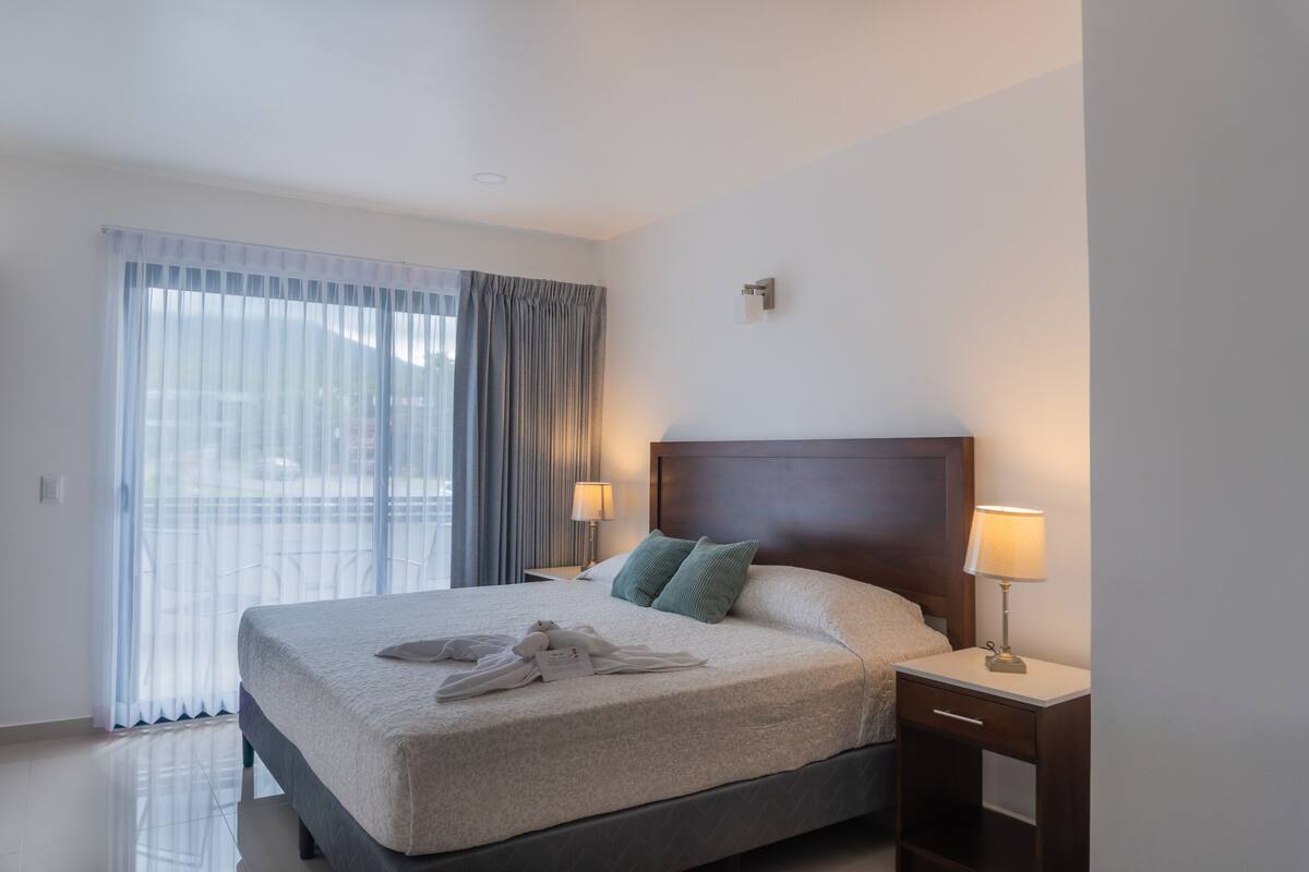Une chambre d’hôtel avec un grand lit, deux lampes de chevet, une table de chevet et une vue dégagée à travers des rideaux légers.